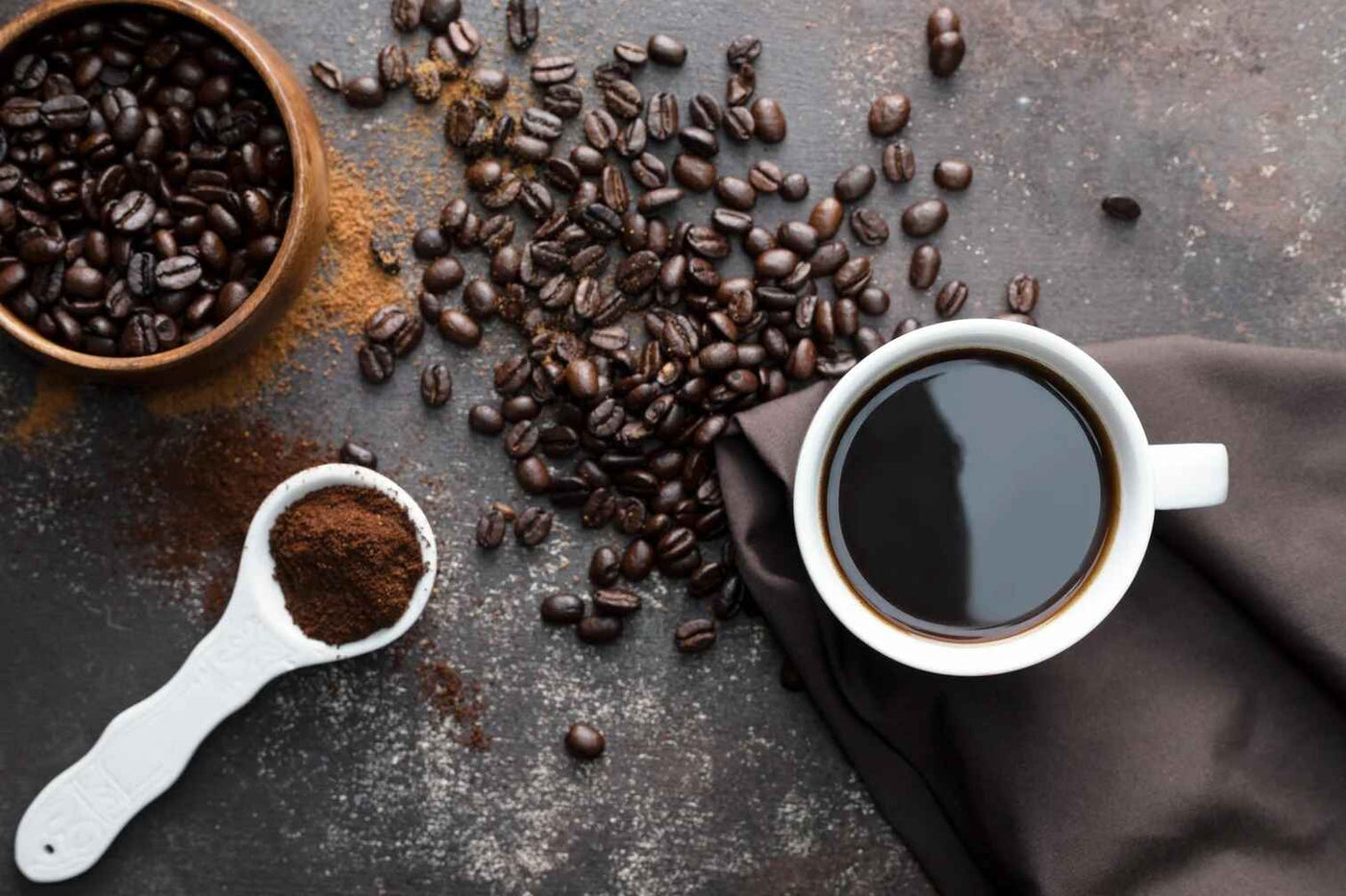 Coffee beans, a cup of coffee, and a scoop of ground coffee on a dark surface.